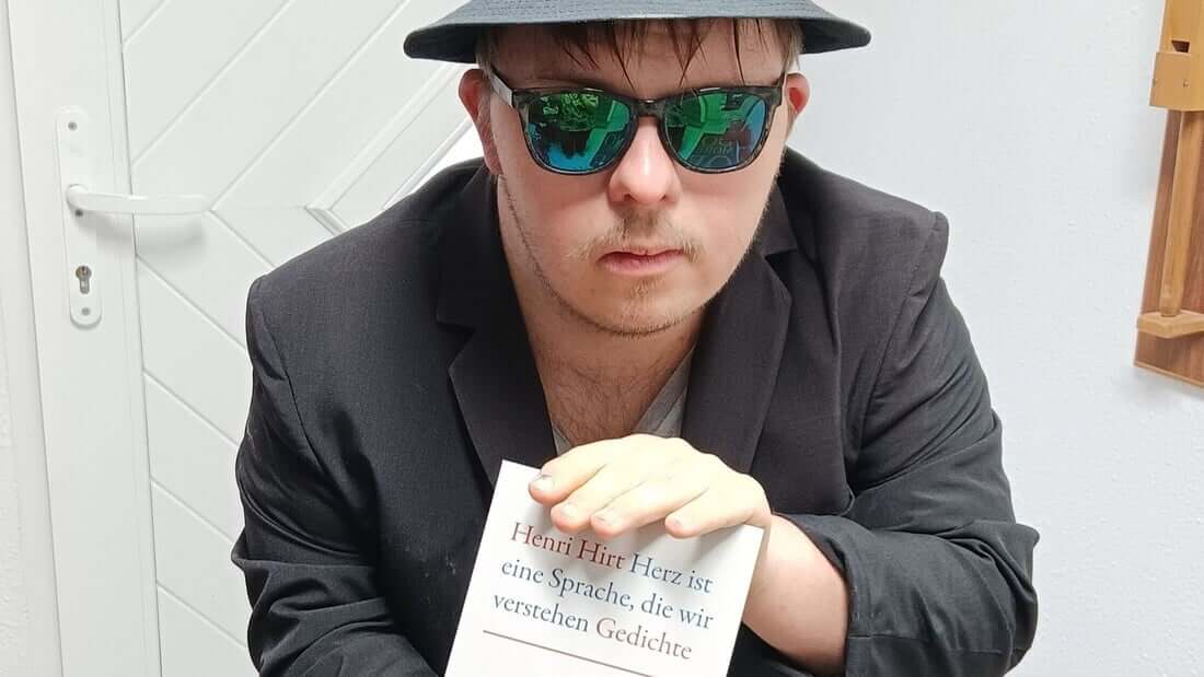 Die ist ein Foto von Henri Hirt. Er trägt einen Hut und eine verspiegelte Sonnenbrille. Er hält sein Buch in die Kamera. Das Buch hat ein weißes Cover und heißt „Herz ist eine Sprache, die wir verstehen